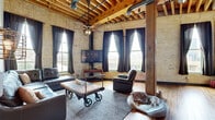 This distinctive loft showcases a compelling industrial aesthetic rooted in its original architectural bones. The living room is a standout space, featuring soaring exposed timber beam ceilings with visible joists, raw ductwork, and original light brick walls that lend an authentic warehouse character. Tall, multi-pane windows flood the room with natural light while offering views of the surrounding urban landscape. The open-plan living, dining, and kitchen area flows seamlessly, anchored by warm hardwood flooring throughout. The kitchen is well-appointed with natural wood cabinetry, granite countertops, stainless steel appliances, and a spacious island with bar seating beneath industrial-style pendant lighting. The dining area continues the industrial theme with exposed timber posts and cage-style pendant lights overhead. The primary bedroom features exposed light brick walls and large windows with dramatic draping, while the second bedroom offers a quieter, more refined atmosphere with neutral tones and carpet underfoot. The bathroom is finished with a rustic-style vanity, dark granite countertops, a large framed mirror, and modern fixtures. Throughout the space, the thoughtful blend of raw, historic materials and contemporary updates creates a cohesive and characterful living environment.