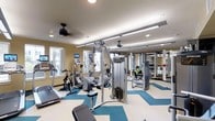 This fitness center offers a comprehensive workout environment designed to accommodate a wide range of exercise needs. The space features a striking teal and cream chevron-patterned floor that adds visual energy to the room, while recessed lighting and overhead fluorescent rail fixtures ensure the area is well-lit throughout. A ceiling fan provides additional air circulation, contributing to a comfortable exercise environment.

The cardio section is well-appointed with treadmills, elliptical trainers, and stationary bikes, several of which are equipped with personal monitors for entertainment or workout tracking. Windows along one wall allow natural light to filter in, creating an open and inviting atmosphere.

Toward the center and rear of the space, a multi-station cable machine serves as a focal point for strength training. The facility extends further to include additional resistance equipment and a dedicated free weights area, with mirrored walls enhancing the sense of depth and allowing users to monitor their form. Wall-mounted televisions are positioned for easy viewing from the cardio machines, adding convenience for longer workout sessions.