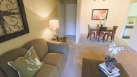 This single-floor space offers a welcoming and functional layout designed for comfortable everyday living. The living area is anchored by a plush sofa accented with decorative throw pillows, complemented by a classic table lamp that casts a warm, ambient glow across the room. Soft, neutral-toned carpeting extends throughout, adding to the cozy and inviting atmosphere.

The open-concept design allows the living area to flow naturally into the dining space, which features a wooden dining table surrounded by upholstered chairs — ideal for gathering and entertaining. A framed floral artwork adorns the wall above the dining area, adding a subtle decorative touch and a pop of color to the otherwise understated palette.

A tiled entryway or hallway connects the main living spaces, providing a practical transition zone and adding visual contrast to the carpeted rooms. Neutral warm-toned walls throughout the space create a cohesive and calming ambiance. The overall design aesthetic leans toward casual comfort, with furnishings and finishes that prioritize livability and warmth. The thoughtful layout maximizes the use of space, making the interior feel both functional and welcoming.
