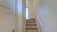 This three-floor space is designed with functional vertical connectivity at its core. The staircase, finished with neutral grey carpeting and a clean white handrail, serves as the primary link between levels and reflects a straightforward, practical design aesthetic. The walls throughout the stairwell are painted in a soft, warm white tone, contributing to a bright and airy atmosphere. A wall-mounted heating unit is visible at the base of the staircase landing, indicating individual climate control built directly into the structure. Natural light filters in through a window visible at the landing, helping to illuminate the transition space between floors. The overall finish quality is consistent and clean, with smooth walls and dark hardwood or laminate flooring visible on the landing area, contrasting with the carpeted stair treads. The narrow, efficient layout of the stairwell suggests a well-organized floor plan designed to maximize usable living space across all three levels. The understated color palette and simple architectural details give the space a neutral, move-in-ready character suitable for a variety of interior styles.