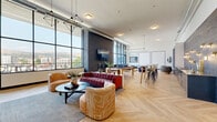 This expansive, single-floor amenity lounge blends industrial character with contemporary elegance across a generous, open-plan layout. Soaring ceilings with exposed ductwork and structural elements lend an urban, loft-inspired aesthetic, while large floor-to-ceiling windows flood the interior with natural light and frame sweeping panoramic views of the surrounding cityscape and hills beyond.

The herringbone-patterned hardwood flooring flows seamlessly throughout the entire space, anchoring the various distinct zones within the room. A relaxed lounge area features a tufted velvet sofa, curved accent chairs, and a round coffee table arranged beneath geometric pendant lighting — ideal for casual gatherings. A statement dark brick accent wall provides a dramatic backdrop for a wall-mounted television, further defining this social hub.

Toward one end, a billiards table anchors a games area, while a long communal high-top table with stylish bar seating serves as a co-working or dining zone. Adjacent to it, a wet bar area is detailed with richly textured wood-paneled walls, warm sconce lighting, and built-in cabinetry with a sink and beverage storage. Whiteboards and additional seating accommodate collaborative or professional use. The space is accessed through sleek glass-paneled double doors, completing its polished, multifunctional design.