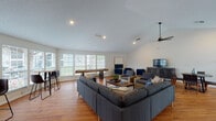 This single-floor space offers a thoughtfully designed open layout that seamlessly connects multiple functional zones, making it well-suited for both social gatherings and relaxed everyday use. Warm hardwood flooring flows consistently throughout, tying each area together with a cohesive, inviting aesthetic.

The main lounge area is generously proportioned, featuring a large sectional sofa paired with wicker accent chairs and a central coffee table. A shuffleboard table adds a recreational element, while a wall-mounted television and media cabinet provide entertainment options. A bank of wide windows with white plantation shutters floods this zone with natural light and offers views of the surrounding greenery.

An adjacent multi-purpose area includes a dining table with chairs, a pair of lounge chairs flanking a side table, and additional bar-height seating — offering flexibility for casual dining, conversation, or small group gatherings. French doors with glass panes connect this area to the exterior, enhancing the sense of openness.

A quieter seating nook, defined by vaulted ceilings and a large framed abstract artwork, features four navy accent chairs arranged around a round side table — ideal for intimate discussions or focused meetings.

Frosted glass sliding and folding partition doors allow the space to be divided into separate zones as needed, providing adaptability. Ceiling fans, recessed lighting, and textured ceilings complete the practical yet comfortable character of this well-appointed space.