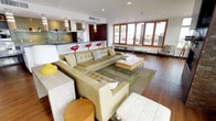 The Sky Lounge at 4040 26th Ave SW in Seattle, WA offers a sophisticated, single-floor communal space designed for both relaxed gatherings and elevated entertaining. The open-plan layout connects a generous lounge area, a full kitchen, and a dedicated dining space, all unified by rich hardwood flooring and a clean, modern aesthetic.

The main lounge is expansive and light-filled, featuring floor-to-ceiling windows and glass doors on multiple walls that invite abundant natural light and frame sweeping outdoor views. A striking brick fireplace anchors one end of the room, topped with a wall-mounted flat-screen display, while sheer white curtains add a soft, airy contrast to the warm wood tones. Folding casement windows open directly to an outdoor terrace, seamlessly blending indoor and outdoor living.

The kitchen is sleek and fully equipped, showcasing green glass subway tile backsplash, granite countertops, stainless steel appliances, and dark cabinetry. A bar counter with bold red barstools connects the kitchen to the lounge, creating a lively social hub.

The dining area features a standout live-edge wood table paired with translucent teal chairs, illuminated by a sculptural ribbon chandelier. A textured accent wall completes this visually dynamic space. Together, these elements make the Sky Lounge a distinctive and functional venue in the heart of Seattle.