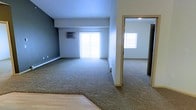 This single-floor residential space offers a practical and comfortable layout designed for everyday living. The main living area is generously proportioned, finished with plush wall-to-wall carpeting that extends seamlessly into the adjoining room, creating a cohesive and unified feel throughout. Neutral-toned walls — including a cool gray accent wall — contribute to a calm and understated ambiance.

Natural light plays a significant role in this space, with large sliding windows in the living area and a secondary window visible in the adjacent room, both allowing bright, diffused light to fill the interiors. A wall-mounted air conditioning unit provides climate control, while baseboard heaters line the perimeter walls, ensuring year-round comfort.

The open sightlines between the main living area and the connected room enhance the sense of space, making the layout feel expansive and airy. A partially visible entryway with hardwood-style flooring offers a clean transition point into the carpeted zones. The overall design is straightforward and functional, with a neutral palette and consistent finishes that provide a versatile backdrop adaptable to a variety of furnishing styles and personal tastes.