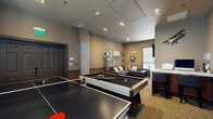 This single-floor recreational space is thoughtfully designed to serve as a social hub for entertainment and relaxation. The main area is generously proportioned, accommodating both a full-size pool table and a ping pong table side by side, providing ample room to move and play comfortably. Track lighting runs along the ceiling, ensuring the gaming area is well-lit, while recessed lighting contributes to a balanced overall ambiance throughout the space.

A dedicated lounge nook is situated toward one end of the room, furnished with a large dark leather sectional sofa accented with decorative pillows. A sizable flat-screen television is mounted on the wall, making this corner ideal for viewing. A window with blinds allows natural light to filter in, complementing the warm, neutral taupe wall tones.

The main gathering wall features a curated gallery of black-and-white framed cityscape photographs arranged around a prominent "gather" wall sign, reinforcing the communal character of the space. Textured wainscoting adds architectural detail along the lower walls, contrasting with the neutral upper tones. The flooring consists of patterned carpet, adding warmth underfoot. Multiple doorways provide easy access and flow, making this a versatile and inviting space for group activities and socializing.
