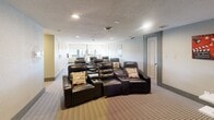 This purpose-built theater room in Fort Wayne, Indiana, delivers a true cinematic experience within a well-appointed, single-floor layout. The main viewing area is anchored by multiple rows of plush black leather recliner seats, each equipped with built-in cup holders, arranged to provide clear sightlines to a large wall-mounted flat-screen display. Recessed lighting and a textured ceiling complement the room's controlled ambiance, while neutral gray walls and patterned carpeting keep the aesthetic clean and focused on the viewing experience. Classic movie posters—including recognizable titles—are displayed throughout, reinforcing the dedicated entertainment atmosphere. A retro-style popcorn machine and concession cart add an authentic theater touch near the screen wall. Adjacent to the main seating area, a high-top table with green accent chairs provides an additional casual gathering space. The room is accessed via a defined entry corridor, giving it a sense of arrival and separation from other areas. Ceiling vents ensure comfortable climate control throughout. Overall, this theater room is thoughtfully designed for group screenings and entertainment, combining functional layout with engaging, movie-inspired décor.