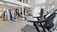 This two-floor fitness center is thoughtfully designed to accommodate a comprehensive range of workout routines in a bright, modern environment. The main floor is anchored by soaring double-height ceilings and floor-to-ceiling windows that flood the space with abundant natural light while offering views of surrounding palm trees and greenery. The open layout houses an impressive array of cardio equipment, including treadmills, stationary bikes, and rowing machines, complemented by a multi-function cable machine and a dedicated power rack for strength training. Stylish pendant light fixtures and clean white walls contribute to a contemporary, motivating ambiance. A metal-railed staircase with an industrial aesthetic leads to the second floor, which offers additional workout space featuring a well-stocked dumbbell rack, a tiered kettlebell station, and specialized resistance machines. Interior windows on the upper level connect visually to the main floor below, maintaining an open and cohesive atmosphere throughout. The lower level also includes a functional exercise area with stability balls, medicine balls, resistance equipment, and direct access via double glass doors. Together, both floors create a fully equipped fitness facility that balances practical functionality with an airy, light-filled design.