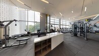 This expansive fitness center is designed with a contemporary architectural aesthetic, distinguished by soaring double-height ceilings and extensive floor-to-ceiling glazing along multiple walls. The generous window coverage floods the interior with natural light while offering open views of the surrounding landscape, creating an energizing workout environment.

The facility is organized into clearly defined zones. A central white countertop island with integrated wood-toned shelving serves as a functional hub, dividing the space and providing storage. The cardio section lines the windowed perimeter, housing a comprehensive row of treadmills, stationary bikes, a stair climber, and rowing machines—all positioned to take advantage of the outdoor views. A dedicated strength training area features a multi-station cable machine, a squat rack with bench, dumbbells arranged on tiered racks, kettlebells, battle ropes, and TRX suspension trainers.

A standout design element is a large-scale artistic mural depicting dynamic athletic figures in bold grey, black, and yellow tones, energizing the strength training zone. Recessed and linear pendant lighting complements the abundant natural light. Rubber flooring throughout provides durability and safety, while perforated ceiling panels and teal accent walls add refined architectural character to the overall space.