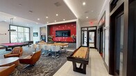 This single-floor amenity space is a dynamic entertainment hub designed with a bold, modern aesthetic. A striking red-and-white color palette runs throughout, anchored by graphic patterned carpeting and crisp white ceilings with recessed lighting. The main game room serves as the centerpiece, housing a red felt pool table, foosball table, shuffleboard table, and a classic pinball machine, alongside a vibrant large-scale artwork that adds artistic energy to the space. A glossy red accent wall frames a large flat-screen television above a built-in bar counter with live-edge wood seating, creating a natural social hub. Adjacent to the game room, a lounge area features a dramatic floor-to-ceiling fireplace clad in cross-sectioned log slices, paired with mid-century-style seating in cognac and red tones. A glass-enclosed private room offers an intimate setting with a poker or game table. Large-scale windows flood the billiards area with natural light and frame views of a lush outdoor pool area with palm trees. The private theater room delivers a cinematic experience with tiered red leather recliner seating, a large projection screen, and bold red and black wall décor. Throughout, the space balances energy and comfort.
