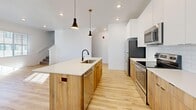 This well-designed two-story home offers 2 bedrooms and 3 bathrooms across two thoughtfully laid-out floors. The main level features an open-concept kitchen and living area anchored by a large central island with a white quartz countertop, an undermount sink, and warm wood-tone cabinetry. White upper cabinets, a subway tile backsplash, stainless steel appliances, and stylish matte black pendant lights complete the modern kitchen aesthetic. Light hardwood flooring flows throughout the main level, complementing the bright, white walls and abundant natural light. The upper floor hosts both bedrooms, including a spacious primary bedroom with vaulted ceilings, plush carpet flooring, and a large window overlooking the neighborhood. The primary bathroom is a standout feature, showcasing a dual-sink vanity with warm wood cabinetry, a white quartz countertop, matte black fixtures, and light tile flooring. A second full bathroom with a tub-shower combination mirrors the same clean, modern design. The carpeted staircase is lined with a large window that floods the space with natural light. Throughout the home, the design palette of white walls, natural wood tones, and matte black accents creates a cohesive, contemporary aesthetic.