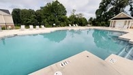 Spring Creek's outdoor pool area is a well-appointed recreational amenity designed for relaxation and leisure. The generously sized swimming pool features clear, blue-green water and a clearly marked shallow end, making it accessible and safe for a variety of swimmers. The expansive pool deck is finished in a light-toned concrete surface, providing ample space for movement and sunbathing. A collection of lounge chairs and seating arrangements are positioned around the perimeter of the deck, offering plenty of spots to rest and unwind. The area is enclosed by a black iron fence, providing both security and a clean, defined boundary for the space. Mature, leafy trees form a dense natural backdrop that surrounds the pool area, creating a sense of privacy and a pleasant natural ambiance. The lush greenery also provides areas of shade and contributes to a calm, retreat-like atmosphere. The single-level layout of the space ensures easy, step-free access throughout the entire pool area. Whether used for recreation or relaxation, this outdoor amenity at Spring Creek offers a comfortable and inviting environment for its visitors.