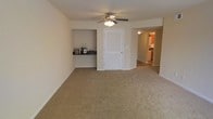 This single-floor space features an open living area finished with wall-to-wall neutral carpeting that contributes to a clean, understated aesthetic. The room is centered beneath a ceiling fan with an integrated light fixture, providing both air circulation and ambient lighting. A distinctive built-in shelving nook is recessed into one wall, offering a dedicated display or storage area. A white louvered door leads to an additional space, possibly a closet or utility area, while a hallway visible in the corner connects to further rooms. Through the hallway, a glimpse of the kitchen reveals cabinetry and tile or hardwood-style flooring, contrasting with the carpeted living space. The walls are finished in a light neutral tone throughout, enhancing the sense of openness. The layout flows naturally between the living area and the connecting spaces, making the overall floor plan feel functional and well-organized. The single-story design ensures easy accessibility throughout.
