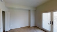 This single-floor space presents a clean, minimalist aesthetic defined by smooth white walls and warm wood-look laminate flooring that runs throughout. The room is open and unfurnished, allowing the full layout to be appreciated. A structural beam detail along the upper wall adds subtle architectural character to the otherwise simple, streamlined interior. Natural light enters through a window fitted with horizontal blinds on the right side of the room, contributing to a bright and airy atmosphere. The neutral color palette — soft white walls paired with light-toned flooring — creates a versatile backdrop well-suited to a variety of uses and furnishing styles. The ceiling is flat and clean, featuring a centrally placed fixture point, and the room benefits from a well-proportioned sense of space. Baseboards provide a neat finish along the floor perimeter. The overall ambiance is calm and understated, with a move-in-ready condition that allows occupants to personalize the space with ease. The combination of natural light, neutral tones, and hard-surface flooring gives the room a practical yet inviting character.