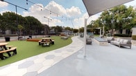 This expansive outdoor amenity area is thoughtfully designed to offer a variety of recreational and social spaces. A curving hexagonal-patterned paver walkway serves as the central spine of the layout, connecting distinct zones and adding a modern geometric aesthetic to the grounds. Lush artificial turf runs alongside the pathway, providing a clean, low-maintenance green surface that hosts multiple picnic tables and contemporary bench seating, ideal for casual gatherings.

A warm-toned wood deck platform anchors one area of the space, furnished with modular lounge seating, umbrella stands, and a built-in grill station, creating an inviting outdoor living and entertaining zone. Overhead string lights are strung throughout the open areas, lending a festive ambiance during evening hours.

A large shade sail canopy covers a separate concrete zone, sheltering a covered game table—likely a pool or ping-pong table—alongside additional lounge seating. A tennis or sport court is visible at the perimeter, enclosed by fencing. Mature trees and landscaped planters frame the space, offering natural privacy and greenery throughout.