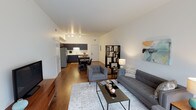 This contemporary single-floor unit showcases a thoughtfully designed open-concept layout that seamlessly integrates the living, dining, and kitchen areas into one cohesive and functional space. Warm hardwood flooring flows throughout, adding a sense of continuity and elegance to the interior.

The living area is well-appointed, offering ample room for comfortable seating arrangements, anchored by a modern sofa and accent chair set in neutral grey tones, complemented by a sleek coffee table and a patterned area rug. A stylish open bookcase serves as a tasteful divider between zones while maintaining the open feel of the space.

The dining area transitions naturally from the living room and provides an intimate setting with a compact dining table and chairs, positioned to benefit from the bright, ambient lighting overhead.

The kitchen features dark cabinetry with a clean, minimalist aesthetic, stainless steel appliances including a full-size refrigerator, and a functional layout designed for everyday use. Under-cabinet and track lighting illuminate the workspace effectively.

Throughout the unit, clean white walls and a neutral palette create a bright and modern ambiance, while the open layout maximizes the sense of space and flow. The overall design is contemporary and move-in ready, appealing to those who appreciate clean lines and functional living.