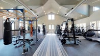 This fitness center is a thoughtfully designed, single-floor facility built to support a comprehensive workout experience. The space is immediately distinguished by its soaring vaulted ceilings, which lend an open and airy atmosphere throughout. Recessed lighting combined with natural light from large windows at the far end of the room ensures the space is bright and inviting at all hours.

The layout is spacious and well-organized, with a central striped runner guiding visitors through the facility. On one side, a power rack with cable attachments and free weight stations is available for strength training, while the opposite side features a cable machine and additional resistance equipment. A multi-functional smith machine and plate-loaded barbell station are also prominently positioned, catering to serious lifters. Large floor-to-ceiling mirrors line the walls, enhancing the sense of space and allowing for proper form checks during exercise.

Cardio equipment and additional strength machines are visible toward the rear of the facility, providing a full range of training options. A mounted flat-screen television is installed near the free weight area for entertainment. The flooring transitions between padded gym matting and striped carpet, defining functional zones within the space. Overall, the fitness center offers a clean, professional environment suited for a wide range of fitness goals.