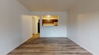 This single-floor unit offers a practical and functional layout across several well-defined areas. The living area is a clean, open space finished with medium-toned wood-style vinyl flooring that runs throughout much of the unit, lending a cohesive and modern appearance. Crisp white walls and white baseboards provide a neutral backdrop throughout.

A standout architectural feature is the pass-through window connecting the living area to the kitchen, allowing visual openness between the two spaces. The kitchen is equipped with dark espresso-toned cabinetry — both upper and lower — paired with light countertops, a single-basin sink, a black range with overhead ventilation, and a full-size black refrigerator. The kitchen also features the same wood-style flooring for easy maintenance.

The bedroom is a well-proportioned room with a window fitted with blinds, allowing natural light to filter in. This room features a distinct multi-toned patchwork carpet, adding visual contrast to the space. A baseboard heater runs along the wall beneath the window.

The bathroom includes a dark-stained vanity with a wide mirror above, a toilet, and a full soaking tub with a shower surround. Light wood-style flooring continues into the bathroom, maintaining consistency with the rest of the unit.