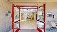 Located at 141 Towpath Dr in Napa, California, this single-story community facility known as Towpath Village presents a thoughtfully designed interior that balances functionality with a warm, inviting atmosphere. The building is accessed through bold red-framed glass double doors flanked by potted olive trees, establishing an immediately welcoming entry. Inside, the lobby features large-format tile flooring, a drop ceiling with recessed panel lighting, and a console table accented with curated artwork — setting a polished tone throughout.

The interior transitions into a lounge and gathering area with warm hardwood flooring, soaring vaulted ceilings, and a striking arched window that frames views of the surrounding landscaped grounds. Dark-stained round tables paired with navy blue upholstered chairs furnish this communal space, which is further complemented by framed artwork on neutral walls.

The fully equipped kitchen showcases rich dark cabinetry, granite countertops, stainless steel appliances including a full-size refrigerator and microwave, a double sink, and a breakfast bar with seating. An adjacent seating nook with comfortable armchairs provides a quiet retreat. The fitness room offers cardio equipment, a round porthole window, and direct outdoor access, completing this versatile and well-maintained community space.