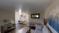 This single-floor space showcases an open-concept layout that seamlessly connects the living and dining areas, creating a cohesive and functional flow throughout. The living area features hardwood-style flooring in a warm, dark tone that extends into the dining space, providing visual continuity across the unit. Neutral-toned walls offer a versatile backdrop, complemented by a textured ceiling that adds subtle architectural interest.

The living area is well-proportioned, accommodating a comfortable seating arrangement centered around a rectangular coffee table, with a rustic wood-framed mirror serving as a decorative focal point on the wall. A bench-style seat with industrial metal legs adds a modern touch to the space.

The dining area, positioned toward the rear of the unit, features a compact table with chairs and benefits from natural light streaming in through vertical-blind-covered sliding glass doors, which also provide access to an outdoor area. A ceiling fan with integrated lighting ensures comfortable air circulation throughout. The overall ambiance is clean, modern, and inviting, with a neutral palette that allows for flexible décor choices.