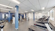Located at 6515 Ridge Ave in Philadelphia's Roxborough neighborhood, this single-floor fitness center offers a generous, open layout designed to accommodate a full range of exercise activities. The expansive exercise room is thoughtfully organized into distinct workout zones, providing ample space for movement throughout. Distinctive blue accent columns punctuate the floor plan, adding a bold visual contrast against the crisp white walls. The durable speckled rubber flooring offers both safety and resilience underfoot, while a drop ceiling with recessed panel lighting ensures the space is brightly and evenly illuminated. A row of windows along one wall allows natural light to filter in, contributing to an open and energizing atmosphere. The cardio zone features multiple treadmills and elliptical machines, while a dedicated strength area is equipped with cable machines, weight benches, free weights, and wall-mounted mirrors. Spinning bikes offer additional variety for cardio workouts. A built-in water fountain provides convenient hydration access. The facility also includes a fully accessible, ADA-compliant bathroom featuring stainless steel grab bars and wood-look vinyl flooring, ensuring the space is welcoming and functional for all users.