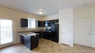 This single-story space includes a well-appointed kitchen that serves as a central feature. The kitchen is outfitted with rich, dark espresso-toned cabinetry that extends to the upper and lower walls, providing ample storage throughout. A suite of black appliances — including a refrigerator, electric range, and built-in microwave — complements the cabinetry for a cohesive, modern aesthetic. Dark granite-style countertops add visual depth and a practical work surface, while a peninsula extension offers additional prep space or casual seating. The sink is positioned beneath a window fitted with horizontal blinds, allowing natural light to filter into the room. The flooring consists of neutral-toned square tiles, lending durability and a clean finish underfoot. The walls are painted in a soft, warm neutral tone, contributing to a bright and open atmosphere. A paneled interior door near the refrigerator provides access to an adjoining area. The overall layout is efficient and thoughtfully designed, maximizing workspace while maintaining an uncluttered, functional environment.