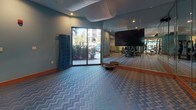 This fitness center is a thoughtfully designed, single-floor facility that blends functionality with stylish interior design. The space is unified by a distinctive blue-and-grey chevron-patterned carpet and warm wood baseboards, complemented by teal-painted walls that create an energizing yet welcoming atmosphere. A decorative draped ceiling element adds a unique architectural touch throughout the main workout area.

The facility is organized into several dedicated zones. A cycling studio features a row of stationary spin bikes arranged on patterned flooring, with geometric-patterned accent walls and full-length mirrors that visually expand the space. Adjacent to this area, a free weights zone houses a comprehensive dumbbell rack, an adjustable weight bench, and built-in wood shelving that displays stability balls and accessories — a practical and organized storage solution.

The strength training section is equipped with a Smith machine and multi-functional cable weight stations, positioned near large windows that invite natural light and views of lush outdoor greenery. Floor-to-ceiling mirrors line multiple walls throughout the facility, reinforcing the sense of openness. Wall-mounted flat-screen televisions are positioned at key areas for entertainment during workouts. The space also includes a stretching and functional training area with balance equipment and a posted workout guide panel, making it a comprehensive and well-rounded fitness destination.