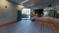 This fitness center is a thoughtfully designed, single-floor facility that blends functionality with stylish interior design. The space is unified by a distinctive blue-and-grey chevron-patterned carpet and warm wood baseboards, complemented by teal-painted walls that create an energizing yet welcoming atmosphere. A decorative draped ceiling element adds a unique architectural touch throughout the main workout area.

The facility is organized into several dedicated zones. A cycling studio features a row of stationary spin bikes arranged on patterned flooring, with geometric-patterned accent walls and full-length mirrors that visually expand the space. Adjacent to this area, a free weights zone houses a comprehensive dumbbell rack, an adjustable weight bench, and built-in wood shelving that displays stability balls and accessories — a practical and organized storage solution.

The strength training section is equipped with a Smith machine and multi-functional cable weight stations, positioned near large windows that invite natural light and views of lush outdoor greenery. Floor-to-ceiling mirrors line multiple walls throughout the facility, reinforcing the sense of openness. Wall-mounted flat-screen televisions are positioned at key areas for entertainment during workouts. The space also includes a stretching and functional training area with balance equipment and a posted workout guide panel, making it a comprehensive and well-rounded fitness destination.