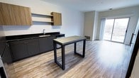This single-floor unit offers a thoughtfully designed open-concept layout where the kitchen and living areas flow seamlessly together. The kitchen is outfitted with dark charcoal cabinetry, dark granite countertops, a stainless steel sink, and a dishwasher. Warm wood-toned upper cabinets and an open shelf add contrast and visual interest. A freestanding dining table with a light wood top and black metal frame anchors the space between the kitchen and the living area, which extends toward a large sliding glass door that floods the interior with natural light and provides access to an outdoor balcony with urban views.

The bedroom features soft carpeting, neutral walls, and a wide window overlooking the surrounding cityscape, creating a calm and airy atmosphere. The bathroom is finished with light tile flooring, a dark-framed vanity with a white sink, a built-in open shelving tower for storage, and a glass-enclosed walk-in shower. Throughout the unit, durable wood-look laminate flooring ties the main living spaces together, contributing to the contemporary and cohesive aesthetic of the overall design.