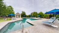 Located at 5200 Greystone Summit Dr in Columbus, Georgia, this outdoor amenity area is centered around a generously sized resort-style swimming pool with crystal-clear blue water and a classic geometric design. The pool deck is finished in attractive stamped concrete, providing ample space for relaxation and socializing. Numerous blue lounge chairs and matching market umbrellas are arranged throughout the deck, offering shaded seating for sunbathers and guests. A separate, smaller wading or leisure pool is positioned adjacent to the main pool, making the area versatile for all types of swimmers. A charming gazebo with a pitched roof and white columns serves as a focal point of the space, providing a shaded gathering area. Brick utility buildings are visible nearby, offering convenient poolside facilities. The entire area is enclosed by a wrought iron fence for safety and privacy, while mature trees and vibrant seasonal plantings create a lush, park-like atmosphere. The surrounding greenery provides a natural backdrop that enhances the serene and inviting character of this well-appointed outdoor recreational space.