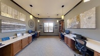 This single-floor business center is thoughtfully designed to provide a functional and inviting workspace. The main room features two built-in workstations positioned along opposite walls, each equipped with a desktop computer, keyboard, and printer, and supported by warm wood cabinetry with ample under-desk storage. Ergonomic task chairs complete each station, ensuring a comfortable work experience. A tall, fully stocked bookcase anchors one corner of the room, offering a curated collection of reading materials. The space is finished with neutral, grey-toned walls accented by bold dark crown molding that frames the room with a polished look. Pendant lighting hangs from the ceiling, providing warm, focused illumination throughout. A central window dressed with a woven valance allows natural light to filter into the room, creating a bright and welcoming atmosphere. Decorative artwork, a large circular wall mirror, and sculptural wall accents add personality and visual interest. A carpeted entry area with additional cabinetry and a workstation serves as a transitional space, with windows overlooking the exterior. The overall ambiance is professional yet comfortable, making this business center a practical and well-equipped resource.