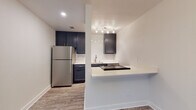 This single-floor space showcases a thoughtfully updated interior with a cohesive, modern aesthetic carried throughout each area. Wood-look plank flooring flows continuously from room to room, providing a warm, unified foundation. The open entryway and living area benefit from natural light through blinded windows, complemented by a decorative chandelier that adds a refined touch.

The kitchen is a standout feature, equipped with dark navy cabinetry, a classic white subway tile backsplash, and stainless steel appliances including a full-size refrigerator. A built-in cooktop is integrated into the breakfast bar peninsula, which also provides additional counter space and a casual dining option. Track lighting above the sink area ensures ample task illumination.

A hallway connects the living spaces to the private areas of the unit, where a closet with paneled double doors provides generous storage. The bathroom is well-appointed with a dark navy vanity topped with a white countertop, a framed mirror, and a tub-shower combination featuring floor-to-ceiling dark tile surround in a vertical layout, creating a sleek, contemporary look. The overall color palette—white walls, dark cabinetry, and neutral flooring—lends the space a clean, modern character.