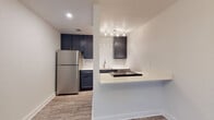 This single-floor space showcases a thoughtfully updated interior with a cohesive, modern aesthetic carried throughout each area. Wood-look plank flooring flows continuously from room to room, providing a warm, unified foundation. The open entryway and living area benefit from natural light through blinded windows, complemented by a decorative chandelier that adds a refined touch.

The kitchen is a standout feature, equipped with dark navy cabinetry, a classic white subway tile backsplash, and stainless steel appliances including a full-size refrigerator. A built-in cooktop is integrated into the breakfast bar peninsula, which also provides additional counter space and a casual dining option. Track lighting above the sink area ensures ample task illumination.

A hallway connects the living spaces to the private areas of the unit, where a closet with paneled double doors provides generous storage. The bathroom is well-appointed with a dark navy vanity topped with a white countertop, a framed mirror, and a tub-shower combination featuring floor-to-ceiling dark tile surround in a vertical layout, creating a sleek, contemporary look. The overall color palette—white walls, dark cabinetry, and neutral flooring—lends the space a clean, modern character.