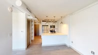 This one-bedroom, one-bathroom unit showcases a contemporary design with an open-concept layout that maximizes the available living space. Light wood-tone flooring runs throughout, creating a cohesive and clean aesthetic. Exposed cylindrical ductwork adds an industrial character to the bright white ceilings, contributing a distinctive urban loft feel.

The kitchen is efficiently designed with white cabinetry, light-toned countertops, and a full suite of stainless steel appliances, including a refrigerator, range, built-in microwave, and dishwasher. A peninsula with an integrated sink serves as both a functional workspace and a casual dining area. Track lighting and pendant fixtures provide focused illumination throughout the space.

The living area benefits from dramatic floor-to-ceiling windows spanning an entire wall, flooding the space with natural light and offering expansive city views. Roller shades provide adjustable light control. A ceiling fan adds practical comfort.

The bedroom continues the light flooring and features a large window with outdoor views, while a built-in shelving unit nearby provides additional storage. The bathroom includes a wide vanity with white cabinetry, a large mirror, and a glass-enclosed shower, all finished in a clean, neutral palette.