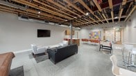 Located at 1335 West Summit Parkway in Spokane, Washington, this single-floor amenity space blends industrial design with modern comfort. The open-concept layout is defined by striking exposed wood beam ceilings with visible ductwork and structural elements, complemented by polished concrete floors that carry throughout the entire space.

The main lounge area is furnished with a tufted leather sofa, white accent chairs, and a coffee table arranged on a patterned area rug, creating an inviting gathering spot. A wall-mounted flat-screen television anchors one end of the lounge, while a large garage-style window floods the adjacent game area with natural light. The game area features a pool table and additional bar-height seating, ideal for social interaction.

A collaborative zone includes a long white communal table with orange chairs alongside a vibrant, colorful wall display that adds a creative focal point to the space. A foosball table nearby reinforces the recreational atmosphere.

The kitchenette is well-appointed with warm wood cabinetry, white countertops, a stainless steel under-counter refrigerator, and a sink — offering practical amenities for residents or guests. An adjacent fitness room, visible through a glass door, provides workout equipment for active use.

Together, these areas form a cohesive, energetic community space that balances functionality with an urban, industrial aesthetic.