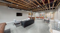 Located at 1335 West Summit Parkway in Spokane, Washington, this single-floor amenity space blends industrial design with modern comfort. The open-concept layout is defined by striking exposed wood beam ceilings with visible ductwork and structural elements, complemented by polished concrete floors that carry throughout the entire space.

The main lounge area is furnished with a tufted leather sofa, white accent chairs, and a coffee table arranged on a patterned area rug, creating an inviting gathering spot. A wall-mounted flat-screen television anchors one end of the lounge, while a large garage-style window floods the adjacent game area with natural light. The game area features a pool table and additional bar-height seating, ideal for social interaction.

A collaborative zone includes a long white communal table with orange chairs alongside a vibrant, colorful wall display that adds a creative focal point to the space. A foosball table nearby reinforces the recreational atmosphere.

The kitchenette is well-appointed with warm wood cabinetry, white countertops, a stainless steel under-counter refrigerator, and a sink — offering practical amenities for residents or guests. An adjacent fitness room, visible through a glass door, provides workout equipment for active use.

Together, these areas form a cohesive, energetic community space that balances functionality with an urban, industrial aesthetic.