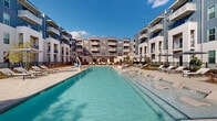 This expansive resort-style pool complex in Vestavia Hills, Alabama, offers a premier outdoor amenity experience set within a modern multi-story residential community. The centerpiece is a generously proportioned swimming pool with crystal-clear turquoise water, designed to evoke a true resort atmosphere. The pool is surrounded by a broad, light-toned paved sun deck that provides ample room for lounging and socializing.

Numerous chaise lounge chairs and sun loungers are arranged throughout the deck, many shaded by large patio umbrellas, offering residents flexible options for relaxation in both sun and shade. Integrated landscaping features raised garden beds filled with native plants, ornamental grasses, young trees, and seasonal shrubbery, adding texture and greenery to the hardscape surroundings. A patch of manicured turf lawn provides an additional casual outdoor gathering area.

A covered pavilion structure at one end of the pool area provides a shaded retreat, complementing the open sun deck. The entire area is enclosed by sleek black metal fencing, ensuring privacy and security. The surrounding multi-story buildings feature contemporary architecture with a combination of blue and white siding, private balconies, and large windows. Under open Alabama skies, this outdoor space delivers a sophisticated, resort-caliber amenity suited for year-round enjoyment.