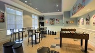 This single-floor game and recreation room offers a well-appointed space designed for social interaction and leisure activities. The room features a tray ceiling with recessed lighting, adding architectural depth and a polished finish to the overall design. Walls are painted in a calming blue-grey tone and accented with white wainscoting panels along the lower half, lending the space a clean, structured aesthetic.

Natural light filters in through large windows dressed with roller shades, brightening the light hardwood flooring that spans the majority of the room. A checkered tile inset near the center of the floor adds a playful design element that complements the recreational atmosphere.

The space is equipped with a foosball table as a central focal point, alongside a set of oversized chess pieces positioned for floor-level play. Bar-height tables paired with ladder-back stools provide seating for casual socializing or spectating. A wall-mounted flat-screen television and a shelving unit offer additional entertainment options. Pop culture and gaming-themed posters decorate the walls, reinforcing the fun, energetic character of the room. Recessed ceiling speakers and a security camera suggest a thoughtfully outfitted communal amenity space.