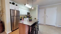 Located at 2991 W School House Ln in Philadelphia, PA, this single-floor residence offers a thoughtfully designed living space with modern finishes and a clean, contemporary aesthetic. The kitchen serves as a central highlight, showcasing stainless steel appliances — including a full-size refrigerator, microwave, and range — set against crisp white cabinetry that extends to the upper walls. Crown molding adds a refined architectural detail throughout, lending an elevated character to the interior. A kitchen island with a neutral quartz-style countertop and warm wood base provides additional prep space and casual seating with bar-height stools, making it ideal for both cooking and entertaining. Pendant lighting hangs above the island, complementing the overall brightness of the space. The flooring transitions from hardwood-style material in the kitchen area to carpet in the adjoining living areas, offering comfort and visual variety. The entryway, visible from the kitchen, features a paneled white door with practical storage nearby. The overall ambiance is light, clean, and inviting, combining functional design with tasteful details that make this Philadelphia residence a comfortable and attractive place to call home.