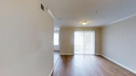 This single-floor residence offers a clean, functional layout with a warm and welcoming ambiance throughout. The main living area is a standout feature, showcasing rich hardwood-style flooring that runs seamlessly across the space, complementing the soft, neutral beige walls and white baseboards that frame each room. A large sliding glass door dressed with vertical blinds serves as a prominent architectural element, flooding the living area with abundant natural light and providing a direct visual connection to the outdoors. An additional window nearby further enhances the bright, airy feel of the space. The textured ceiling adds subtle depth to the interior, while a centrally mounted ceiling light fixture provides warm, consistent illumination. A ceiling-mounted HVAC vent ensures year-round climate comfort. The open sightlines between the living area and the adjacent space create a sense of continuity and ease of movement throughout the floor plan. Crisp white trim and door casings provide clean contrast against the warm wall tones, contributing to a cohesive and well-finished interior aesthetic. Overall, this residence presents a practical and inviting environment suited to comfortable everyday living.