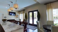 Located at 7331 Skyline Dr in Columbus, Ohio, this single-floor space delivers a polished and sophisticated atmosphere through a carefully curated combination of design elements and high-end finishes. The interior showcases rich dark hardwood flooring that grounds the space with warmth and contrast, while tray ceiling detailing adds architectural depth and visual interest overhead. Elegant pendant lighting, including a classic drum shade fixture, casts a refined glow throughout the open layout. A granite-topped counter anchors one side of the room, complemented by richly upholstered leather barstools with nailhead trim detailing — a hallmark of classic, transitional design. The seating area features a mix of a cream wingback chair and leather club chairs, offering a comfortable and inviting arrangement. Tall, manicured topiary plants flank a set of dark-framed double glass doors, which open to an outdoor area and invite abundant natural light into the interior. Cream drapery panels frame the windows, softening the space and adding to its elegant character. A flat-screen television is mounted on the wall, lending a functional touch to the otherwise refined environment. Overall, the space strikes a confident balance between sophistication and comfort.