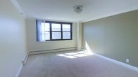 This single-floor space features a spacious open room with a clean, minimalist design. The room is finished with wall-to-wall neutral-toned carpet, which adds warmth underfoot and contributes to a quiet, comfortable atmosphere. The walls are painted in a soft beige tone, creating a bright and airy feel throughout.

A prominent multi-pane window with dark framing dominates one wall, allowing generous amounts of natural light to flood the interior. The window offers views of the surrounding exterior, and sunlight casts distinct rectangular patterns across the carpet, enhancing the sense of openness. Sheer white curtains are present, providing the option for diffused light or added privacy.

Baseboard heating runs along the lower portion of the walls, indicating a practical and efficient heating system well-suited to the layout. The ceiling is smooth and flat, fitted with a single flush-mount light fixture centrally positioned to provide even overhead illumination. Crown molding details add a subtle architectural finish to the room.

Overall, the space presents a functional and well-lit environment with straightforward finishes, making it highly adaptable for a variety of uses.
