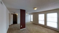This single-floor space offers a straightforward and functional layout designed for comfortable everyday living. The main living area is a standout feature, characterized by plush carpet flooring and an open feel enhanced by multiple windows fitted with white blinds that allow natural light to filter throughout the room. A bold, deep red accent wall serves as a striking architectural focal point, adding a touch of color contrast against the neutral beige walls and textured ceiling. Ceiling fans and flush-mount light fixtures provide both practical illumination and airflow throughout the space. The textured popcorn ceiling adds a classic residential character to the interior. A hallway visible from the main living area leads toward additional rooms, hinting at a well-organized floor plan. The entry area, partially visible in the background, connects seamlessly to the rest of the unit. Overall, the space presents a clean, move-in-ready environment with a warm, neutral palette punctuated by the dramatic accent wall, making it a practical and visually interesting place to call home.