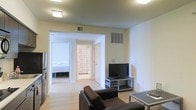 This single-floor residential space offers a straightforward and functional layout designed for comfortable everyday living. The living area serves as the central hub of the home, featuring light hardwood flooring that runs throughout and clean, neutral-toned walls that create a bright and welcoming atmosphere. Flush-mount ceiling lighting provides consistent illumination across the space, complementing the natural and artificial light balance. The living room is furnished with a dark leather sofa and a flat-screen television set on a sleek media stand, establishing a practical and comfortable entertainment area. A glass-top coffee table adds a modern touch to the seating arrangement. From the living area, an open doorway leads directly into a bedroom, which is visible in the background and appears to offer a calm, restful environment. A decorative patterned door nearby adds a subtle design detail to the corridor area. Ventilation grilles are integrated into the walls and ceiling, indicating a central HVAC system for climate control throughout the unit. The overall aesthetic is minimal and clean, making the space versatile and easy to personalize. The single-floor plan ensures seamless flow between areas, making it a practical and well-organized living environment.
