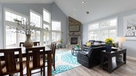 This single-floor space makes a strong architectural statement with its dramatic vaulted ceiling, which draws the eye upward and creates an expansive, airy atmosphere throughout the open-concept layout. The walls are finished in a cool, neutral gray-blue tone that complements the natural light pouring in from the numerous large, white-trimmed windows positioned along multiple walls, offering views of the surrounding outdoor environment.

The focal point of the main living area is a floor-to-ceiling natural stone fireplace, constructed from varied river rock and extending to the peak of the vaulted ceiling. A flat-screen television is mounted directly above the fireplace, making it both a functional and decorative centerpiece. The seating area features dark leather reclining sofas arranged around a circular coffee table, anchored by a bold teal and white patterned area rug that adds a vibrant contrast to the light hardwood flooring beneath.

Adjacent to the living area, a dining space includes a dark wood high-top table with matching bar-height chairs, providing a casual yet defined eating area. Decorative greenery and tasteful accents are placed throughout, contributing to the welcoming and well-curated ambiance of the space.