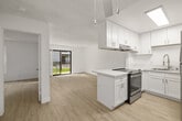 This single-floor unit offers a thoughtfully arranged layout encompassing one bedroom, one bathroom, and a combined kitchen and living area. The open-plan living and kitchen space is bright and airy, featuring light wood laminate flooring and a textured ceiling throughout. Large sliding glass doors in the living area lead to a private balcony that overlooks a well-maintained courtyard with lush green landscaping, palm trees, and a central water feature — providing a pleasant outdoor outlook.

The kitchen is efficiently designed in a U-shaped configuration with white shaker-style cabinetry, light countertops, stainless steel appliances including a refrigerator, dishwasher, and range, plus a skylight that introduces additional natural light. A peninsula with a built-in cooktop and overhead exhaust fan connects the kitchen to the living space. A ceiling fan is also present in this shared area.

The bedroom is a comfortable, sunlit room with a window framing views of the surrounding greenery, light wood flooring, and clean white walls. The bathroom features a full bathtub with shower, toilet, and wood-look flooring in a clean, white finish. The overall aesthetic is clean, modern, and minimalist.
