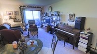 This single-floor space exudes a cozy, lived-in warmth defined by its eclectic and personal character. The main living area serves as the heart of the home, thoughtfully arranged to accommodate both relaxation and entertainment. A classic upright piano anchors one side of the room, lending a musical and cultural touch to the space. Opposite, a well-stocked shelving unit displays a curated collection of personal mementos, decorative items, and media equipment, adding depth and personality to the walls. Seating is both stylish and inviting, with an ornate wrought-iron and glass coffee table at the center and an elegant wingback armchair nearby, creating a refined yet comfortable gathering area. Soft, plush carpeting runs throughout, contributing to the room's warm ambiance. A sliding glass door along one wall floods the space with natural light, while decorative garland framing the doorway adds a festive, homey touch. Framed portraits and artwork adorn the walls, reinforcing the personal and welcoming atmosphere. The overall design reflects a thoughtfully curated space rich in character, blending functional living with artistic sensibility.