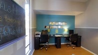 This single-floor business center offers a functional and visually engaging workspace designed to support productivity. The room features two dark-finished workstations, each equipped with a desktop computer and complemented by stylish green desk lamps that provide focused task lighting. A multifunction printer is conveniently positioned at one end of the workspace, making document management straightforward for users.

The interior design draws the eye with a bold teal accent wall that serves as a striking focal point, adorned with decorative wave-shaped mirrors that add depth and a modern aesthetic to the space. The lower portion of the walls features a neutral gray tone, creating a balanced, two-tone color scheme. Warm hardwood-style flooring adds a welcoming contrast to the contemporary furnishings.

Ergonomic rolling chairs in black upholstery are provided at each workstation, ensuring comfort during extended use. Natural light filters in through a large window along the adjacent wall, brightening the space and reducing reliance on artificial lighting. The high ceiling height further enhances the open, airy atmosphere of this well-appointed business center.