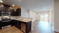 This thoughtfully designed single-floor residence offers a clean, modern aesthetic with high-quality finishes throughout. The open-concept kitchen and living area form the heart of the home, anchored by a peninsula with light stone countertops that seamlessly connects the two spaces. The kitchen is outfitted with rich espresso cabinetry, a white subway tile backsplash, stainless steel appliances including a gas range, over-the-range microwave, dishwasher, and refrigerator. Warm-toned hardwood-style flooring runs throughout the main living areas, complemented by recessed lighting and crisp white walls that create a bright, welcoming atmosphere. Sliding glass doors in the living area open to an outdoor balcony, allowing natural light to pour in. The bedrooms feature plush carpeting and generously sized windows that frame outdoor views, providing a comfortable, serene retreat. Two bathrooms serve the unit: one with a walk-in glass-door shower and dark vanity cabinetry, and another with a full soaking tub and shower combination, both finished with neutral tile flooring and light stone countertops. The overall design balances contemporary style with functional living spaces, offering a well-appointed, move-in-ready environment.
