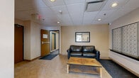 Located at 7301 Rue Beaubien E in Anjou, Québec, this single-floor amenity space provides residents with a functional and practical set of shared facilities. The layout encompasses a lobby/hallway area, a dedicated gym, and additional support rooms.

The entry lobby sets a welcoming tone with tile flooring in a herringbone pattern, recessed lighting within a drop-panel ceiling, and warm neutral wall tones. A black leather loveseat and a wooden coffee table offer a comfortable waiting area, while a built-in wall of individual mailboxes serves as a practical mail collection point. An elevator and clearly marked exits ensure easy access throughout the building.

The fitness room is generously proportioned and well-lit by both recessed and fluorescent panel lighting. Carpeted flooring spans the workout area, which is outfitted with a broad selection of equipment including a treadmill, elliptical trainer, stationary bikes, a multi-station cable machine, weight benches, free weights, and a Precor rowing machine. Windows along one wall introduce natural light, enhancing the open, airy atmosphere of the space. The clean white walls and uncluttered layout make for a focused and functional workout environment suitable for a variety of fitness routines.