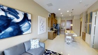 This single-floor unit offers a thoughtfully designed open-concept layout that seamlessly connects the living, dining, and kitchen areas into one cohesive, functional space. The living area features a comfortable grey sofa accented with decorative pillows and a bold navy geometric area rug, complemented by two striking large-format abstract artworks that add a contemporary flair to the neutral warm-beige walls. Recessed lighting throughout the ceiling ensures the space is well-lit and inviting.

The kitchen is efficiently arranged with warm-toned wood cabinetry that extends to the upper walls, providing generous storage. Stainless steel appliances, including a refrigerator and range, lend a modern, polished look. A breakfast bar with a light-toned stone countertop and sleek bar stools bridges the kitchen and living area, creating an ideal casual dining and entertaining space. Pendant lighting above the bar adds both functionality and a design accent. Light-toned large-format floor tiles run throughout the entire unit, creating a clean, continuous aesthetic that enhances the sense of openness and ease of movement across the space.